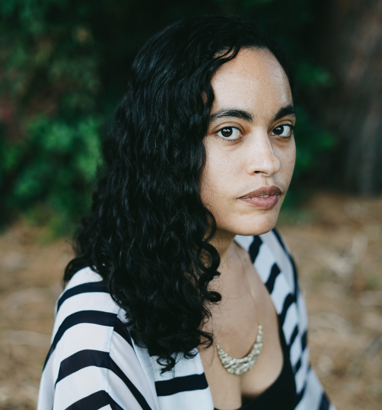 Woman of medium-light skin with long black hair in striped cardigan seated, in background is a natural setting