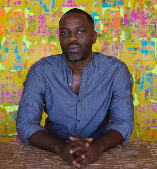 A man of dark skin tone in a blue shirt sits with hands clasped in front of a brightly colored wall, predominantly yellow with patterned blocks of orange, green, pink, and red