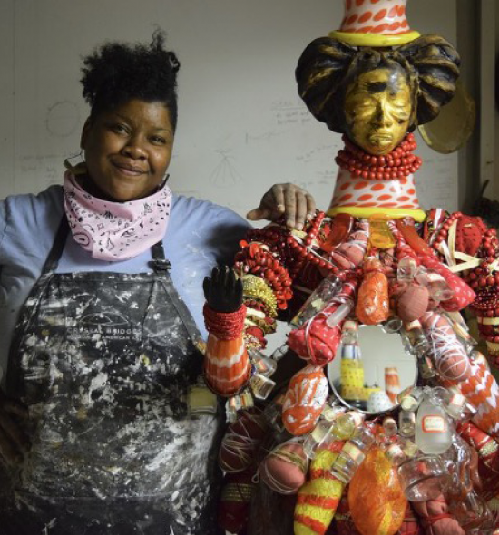 Woman of dark skin tone in a spattered apron, blue shirt, and pink handkerchief tied around her neck stands with a hand resting on the "shoulder" of a sculptural figure, composed of red beads, bronze, cloth, and glass bottles