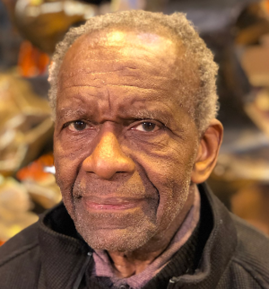 An elderly man of dark skin tone seen from the shoulders up in a black jacket and shirt, in the background, out of focus is a jumble of objects