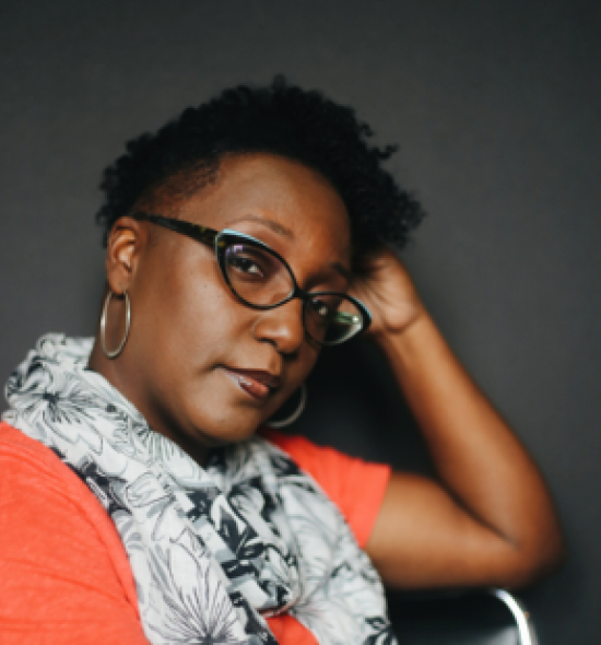 Woman of dark skin tone with short hair and glass, in orange shirt and white scar with black pattern, seen from shoulders up, rests head on left hand, against a black background