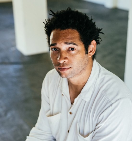 Man of medium skin tone in a white shirt sits in a space with a concrete floor with white square pillars