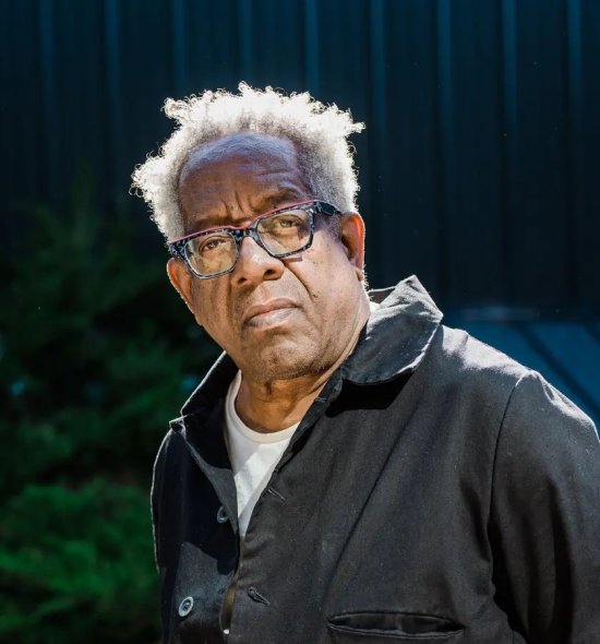 A man of dark skin tone with white-gray hair and glasses in a dark gray jumpsuit stares into the camera lens