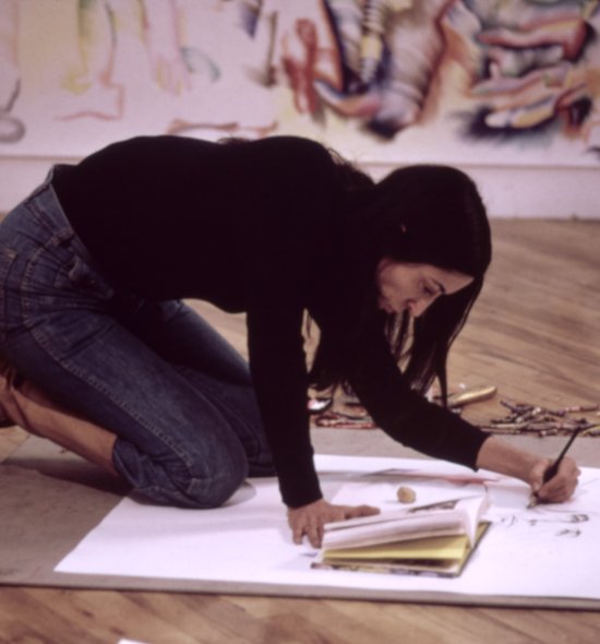 A woman with dark hair leaning over a sketchpad and drawing while on the floor
