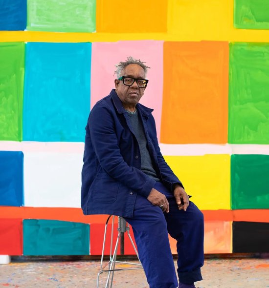 Portait photo of an older man with darker skin tone and grey hair sitting on a stool in front of a large vibrant geometric/grid like painting