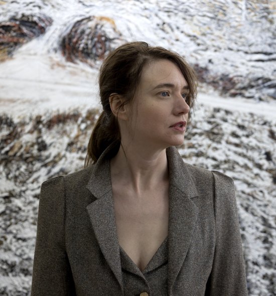 Photo of a woman with short dark hair, fair skin, wearing a blazer, standing in front of a textured painting