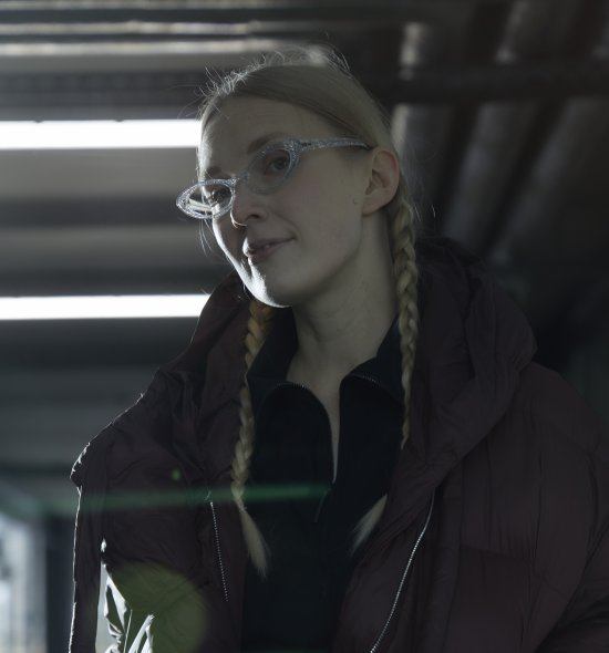 Darkly lit image of a woman with blonde braided hair, light skin tone and glasses wearing a black zip up coat