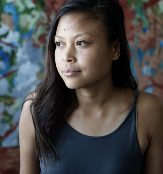 Headshot of a woman with long dark hair, brown eyes, medium skin tone, wearing a blue tank top in front of a painting 
