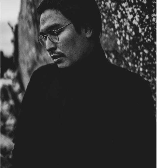 Black and white image of a man leaning against a wall looking down with dark hair and glasses, wearing a black turtleneck
