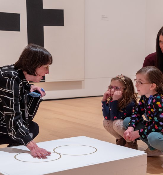 A person kneeling with her hand on a stack of papers talking to two small children and an adult