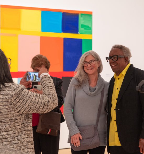 A woman taking photo of a woman with grey hair and light skin tone with a man in a yellow shirt and black jacket with dark skin tone and glasses in an art gallery