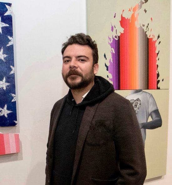 A person with dark hair and facial hair wearing a dark hoodie and blazer, standing in front of vibrant paintings