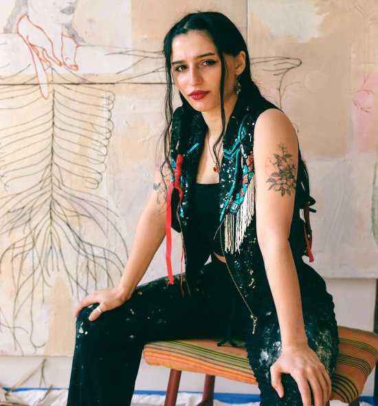 A person sitting on a stool with their hands resting on the tops of their legs, looking seriously into the camera