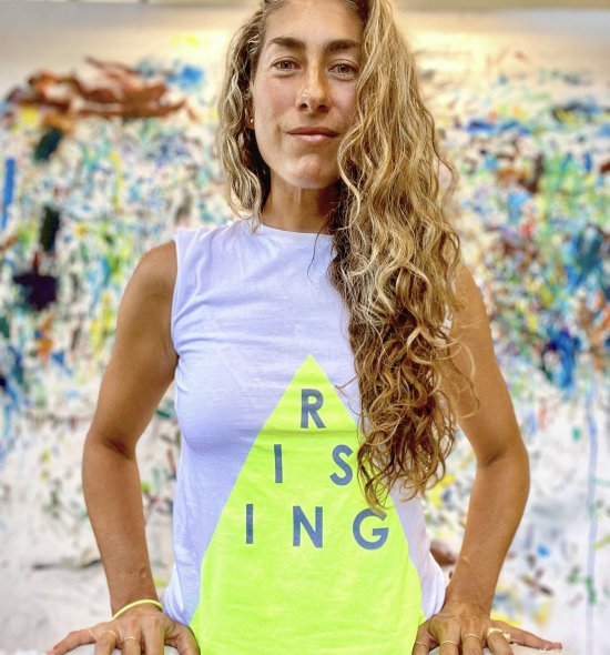 A person with long blonde curly hair wearing a grey tank top with a yellow triangle with the word 'RISING' written on it
