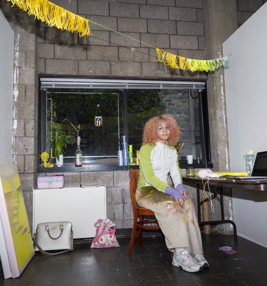 A person with curly fair hair sitting in a studio with pops of yellow decor and art throughout