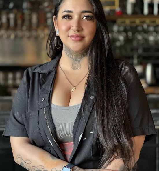 A person with long dark hair and tattoos on their neck and hand and a nose piercing