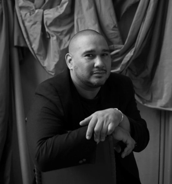 Black and white photo of a person in a dark suit posing in front of draped fabrics background