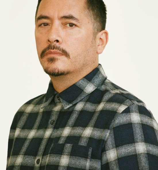 Headshot of a person with dark facial hair wearing a plaid long sleeve button down, standing in front of a white wall