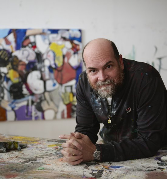 A person with grey facial hair, leaning on an artist studio with a vibrant painting in the background