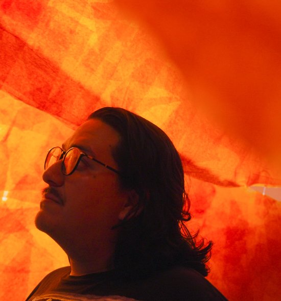 A person with dark hair and glasses under an orange transparent fabric 
