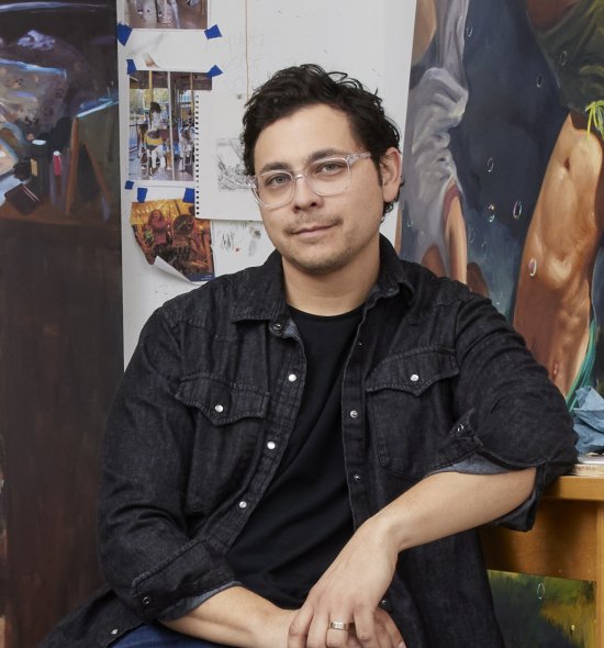 A person with dark hair and clear framed glasses and dark shirt sitting in front of an art studio setup