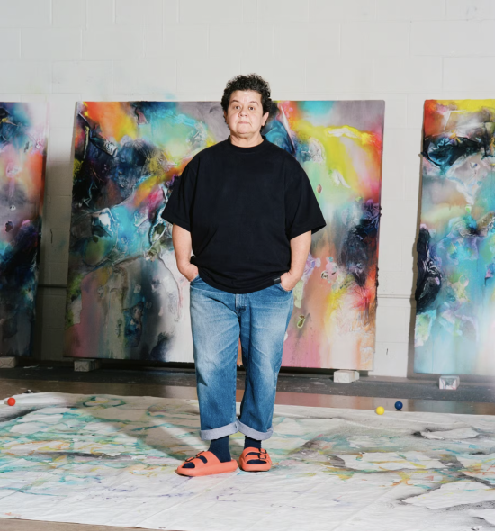 A person in a black tee shirt and denim jeans and red sandals stands in front of large vibrant abstract paintings