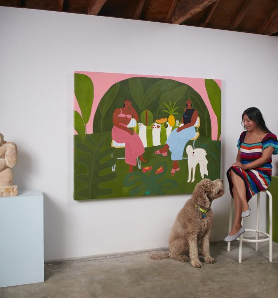 A person in a striped dress, sitting on a stool and petting a large labradoodle in front of a painting of a garden party