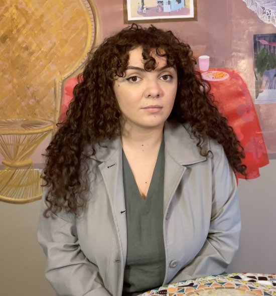 A person with long, dark, curly hair wearing a green shirt and grey coat