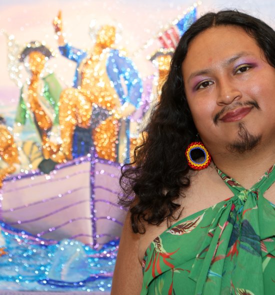 A person with rainbow earrings and green floral shirt standing in front of a shimmery painting of people in a boat