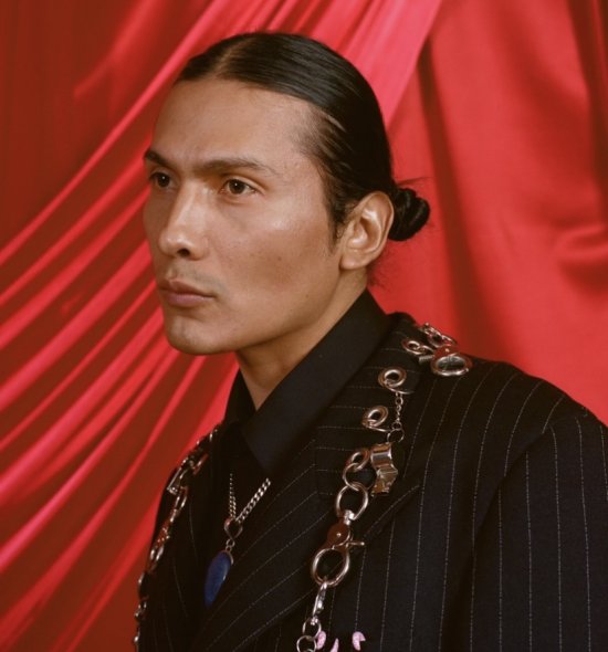 A person with dark hair pulled into a slick bun, wearing a black pin striped suit, looking to the left, in front of a red draped backdrop