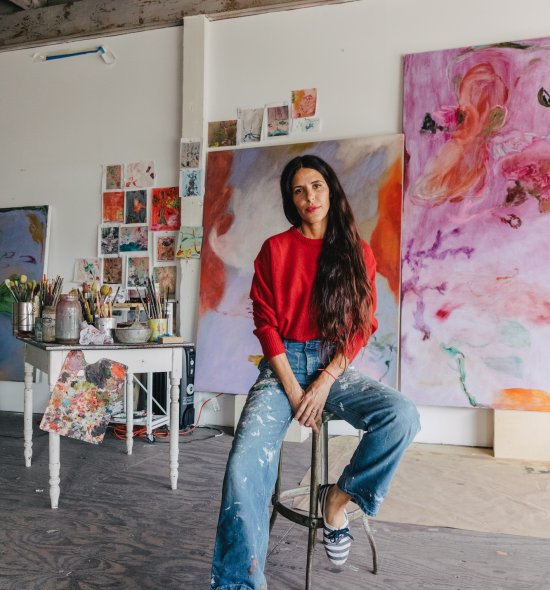 A person in a red sweater and jeans, sitting on a stool in an art studio with large vibrant abstract works in the background