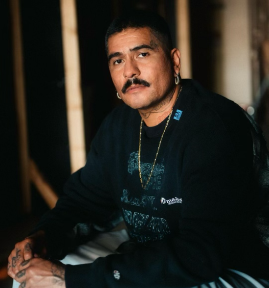 A person with dark hair and a dark mustache and tattoos on the side of their forehead and neck, wearing a long sleeve black shirt and gold jewelry