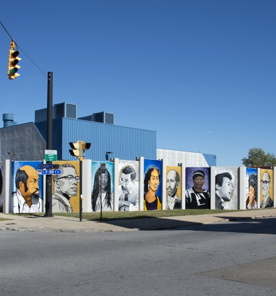 A mural featuring portraits of 28 people on a paneled wall that stretches around a city corner