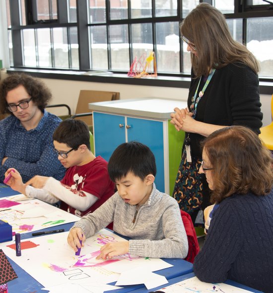 Two kids creating art with two adults seated next to them and one adult standing behind to guide them