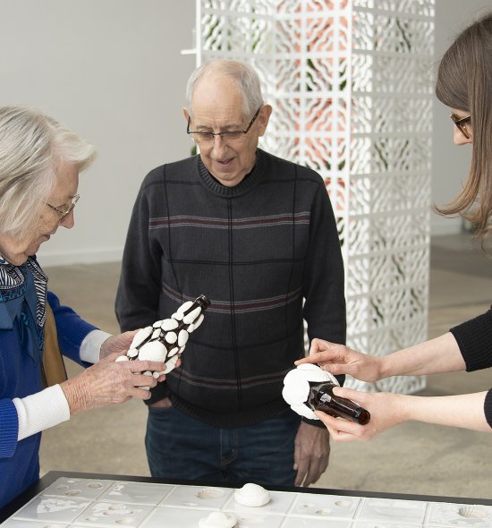 An older woman holds a glass bottle with clay seashells on it while an older man and younger woman look on