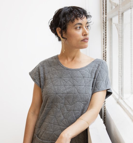 Headshot of a woman with short dark hair, medium skin tone, wearing a grey shirt, leaning against a railing looking out a window