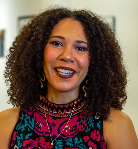 Woman of medium-light skin with curly brown hair in a brightly green and magenta sleeveless top seen from the shoulders up, a row of paintings hangs on the wall behind her