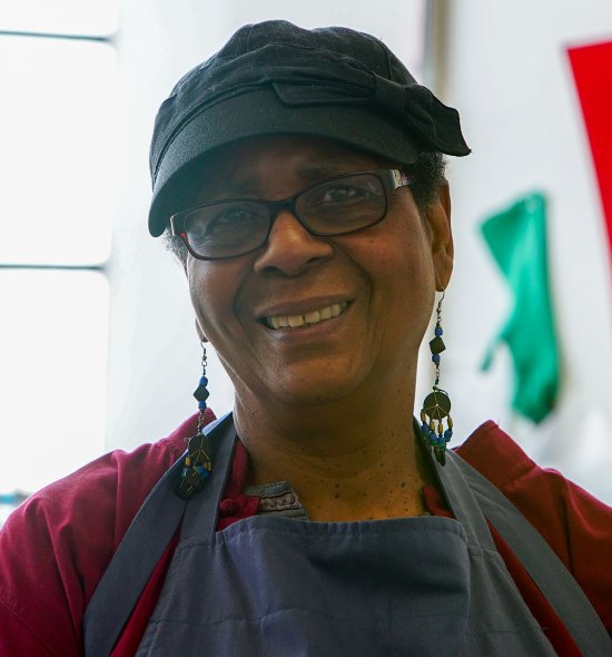 A woman of dark skin tone with a black hat, glasses, and long beaded earrings wearing a dark red shirt and blue apron with a large window and red print behind her